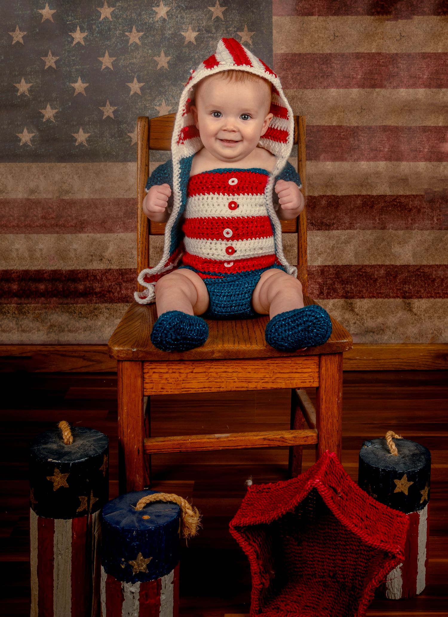 Baby in patriotic red, white, and blue crochet outfit sits on wooden chair before American flag backdrop.
