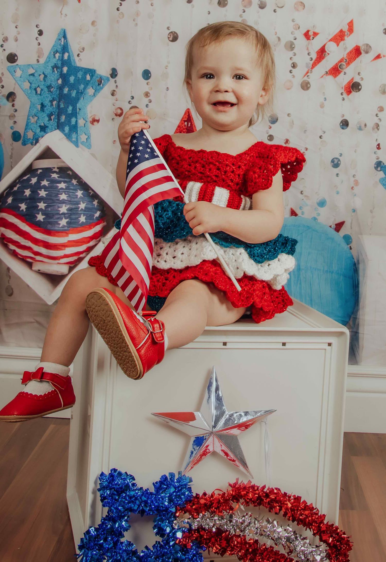 Smiling baby girl in red crochet dress holding American flag, sitting on white box with patriotic 4th of July decorations.