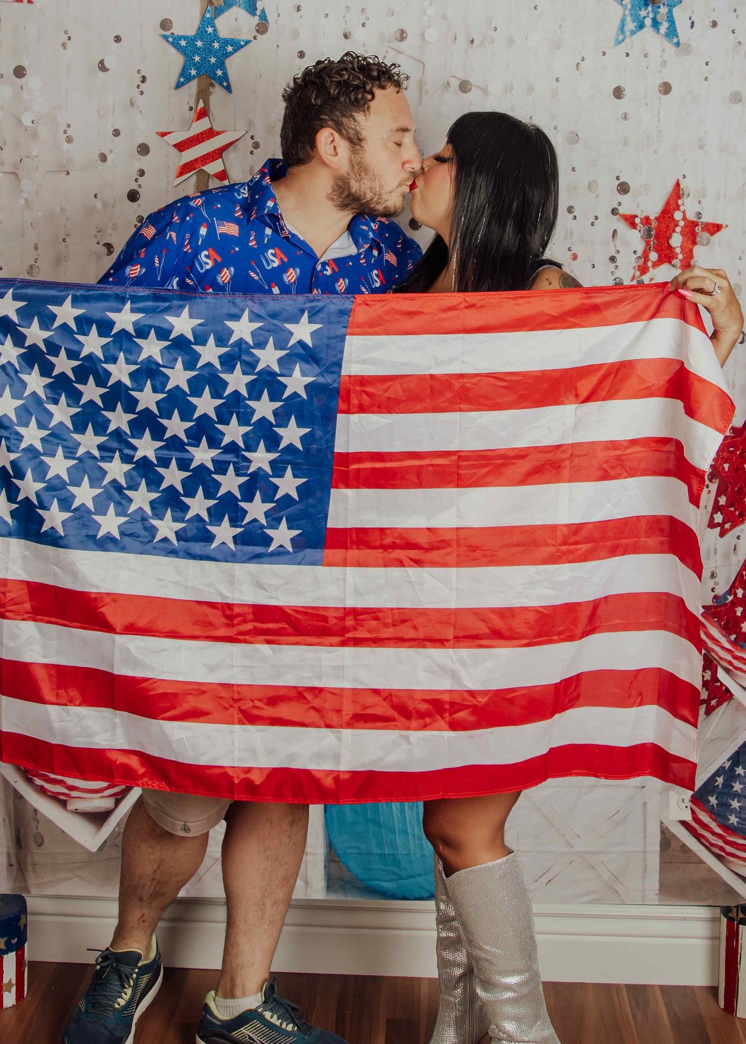 Couple kissing behind a large American flag, wearing patriotic outfits, with red, white, and blue decorations in the background.