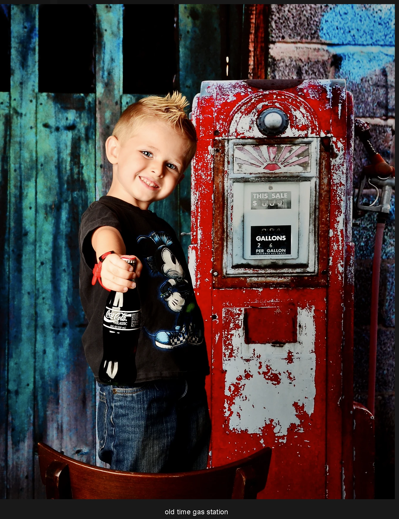 Young boy smiling beside a vintage red gas pump at an old-time gas station, wearing a black t-shirt.