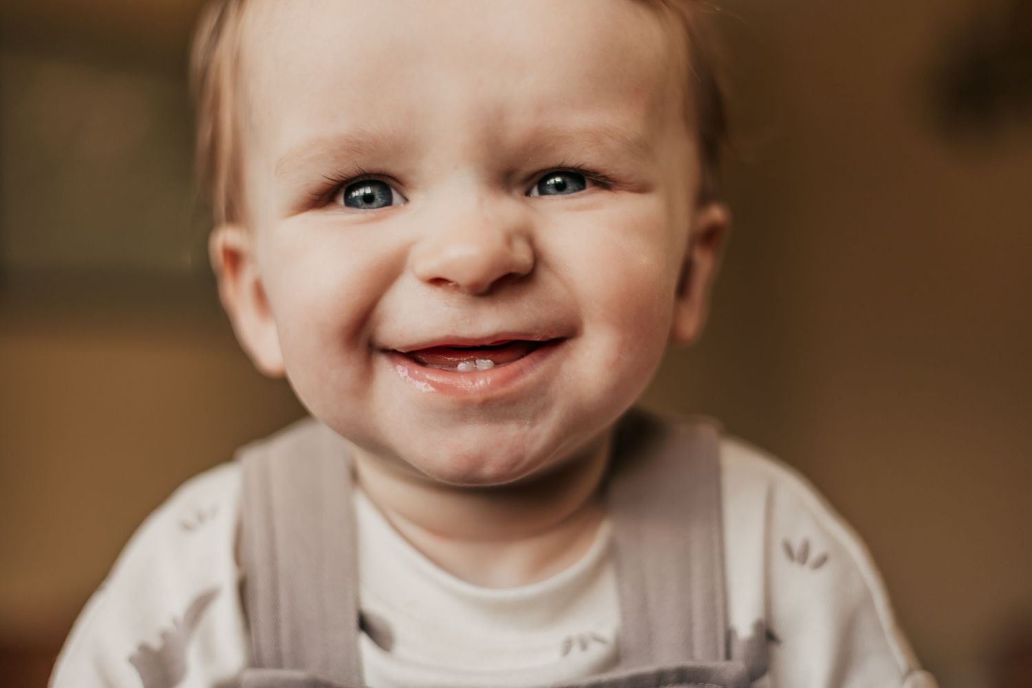 Adorable smiling baby with blue eyes and tiny teeth wearing a gray outfit in a warm indoor setting |Professional Milestone Sessions for Baby