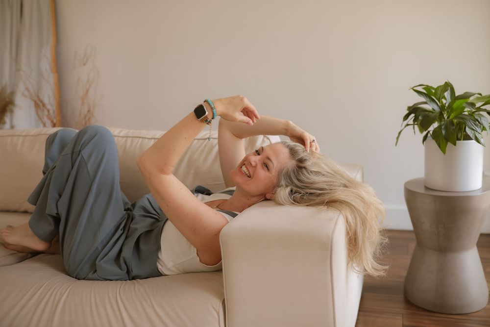 Woman relaxing on beige sofa wearing smartwatch, lying down in bright modern living room with indoor plant.