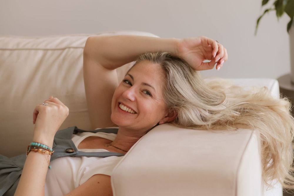 Smiling middle-aged woman with blonde hair relaxing on a cream sofa, raising her arm joyfully indoors.