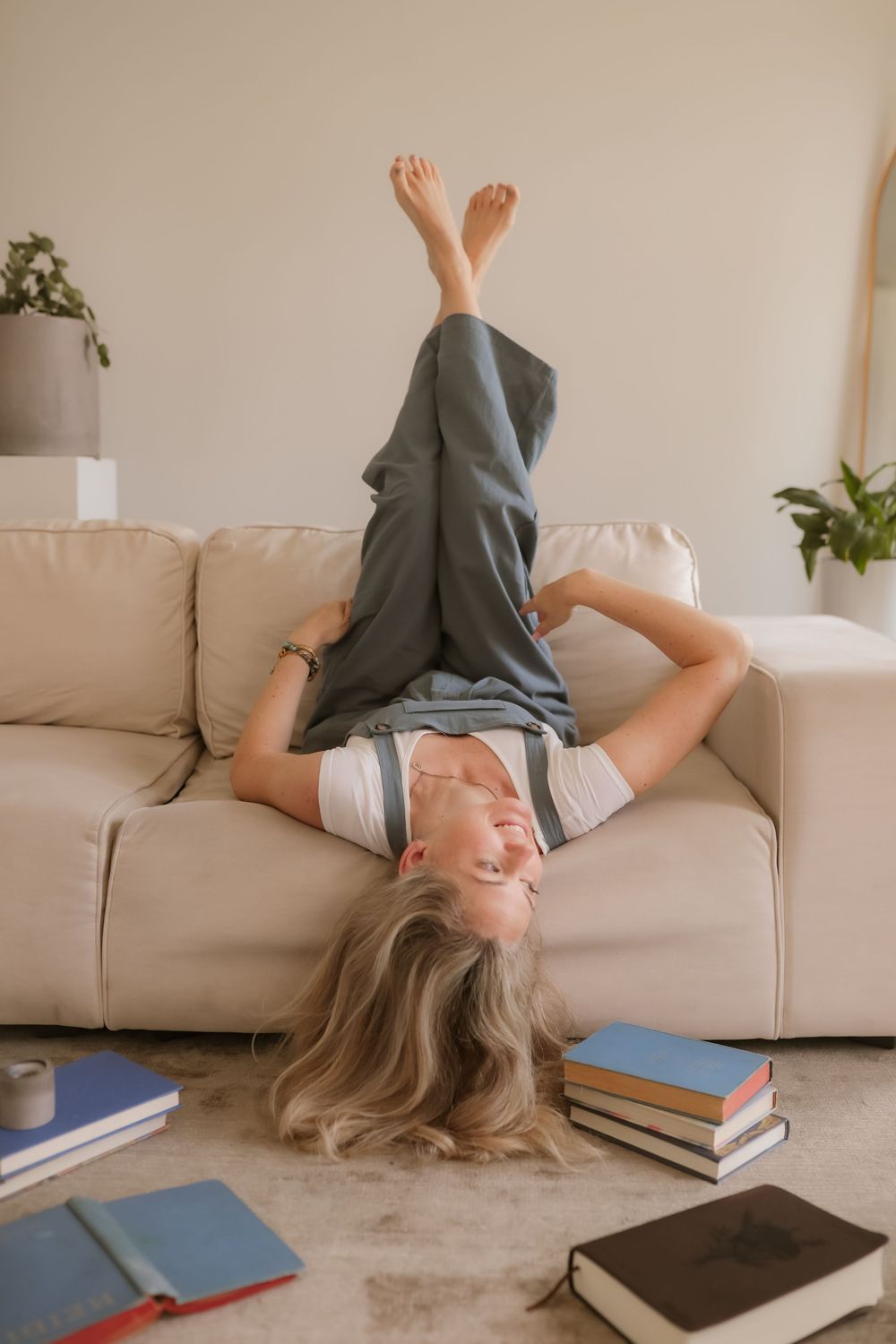 Young woman lying upside down on a beige sofa, smiling, with books scattered on the floor around her.