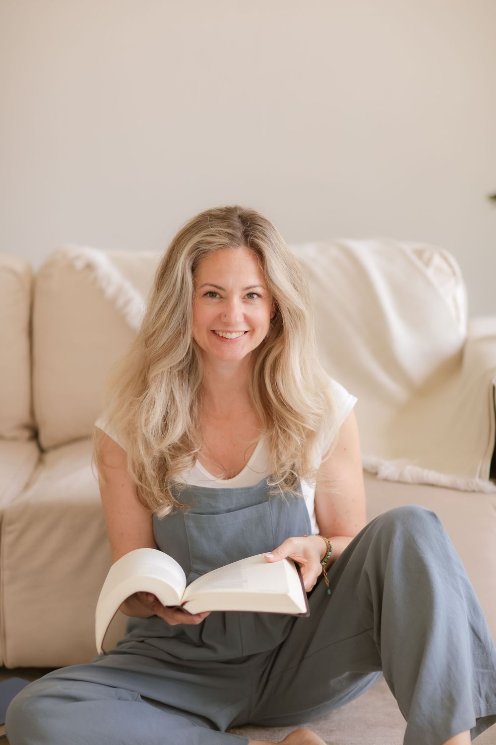 Smiling blonde woman in gray overalls sitting on floor holding open book near cream sofa in bright living room.