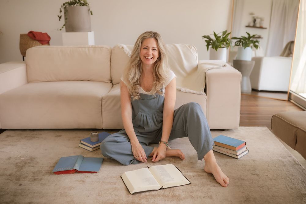 Smiling blonde woman sitting cross-legged on a rug surrounded by books in a bright, cozy living room.