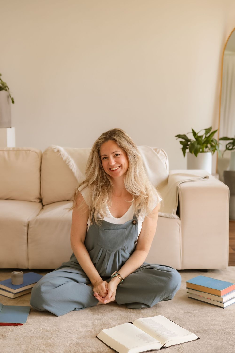 Smiling blonde woman sitting cross-legged on floor surrounded by books near a beige sofa in a cozy living room.