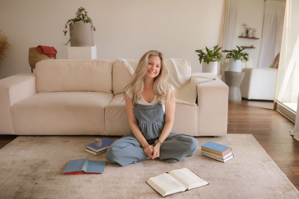 Smiling blonde woman sitting cross-legged on carpet surrounded by books near a white sofa in a bright living room.