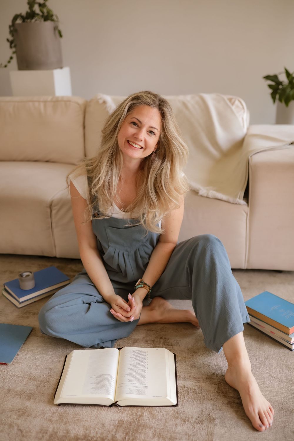 Smiling blonde woman sitting cross-legged on floor with open book, surrounded by stacked books near a beige sofa.