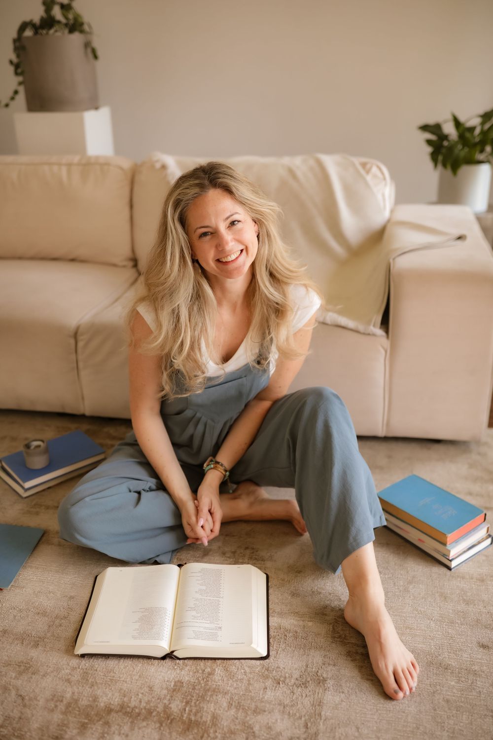 Smiling blonde woman sitting cross-legged on floor with open book, surrounded by stacked books near a couch.