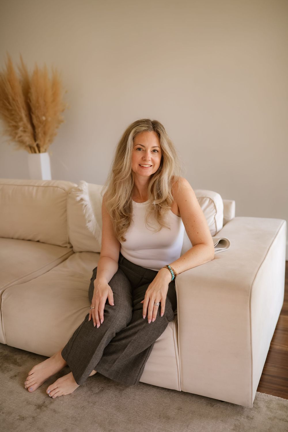 Smiling blonde woman in white tank top and dark pants sitting on beige sofa in modern living room.