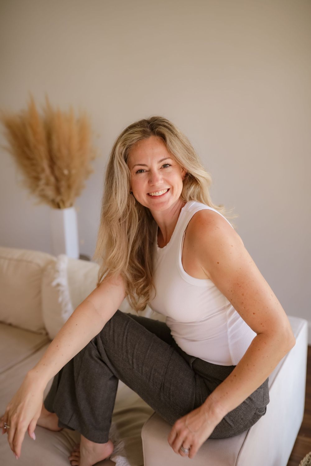 Smiling blonde woman in white tank top and gray pants sitting relaxed on a cream sofa indoors.