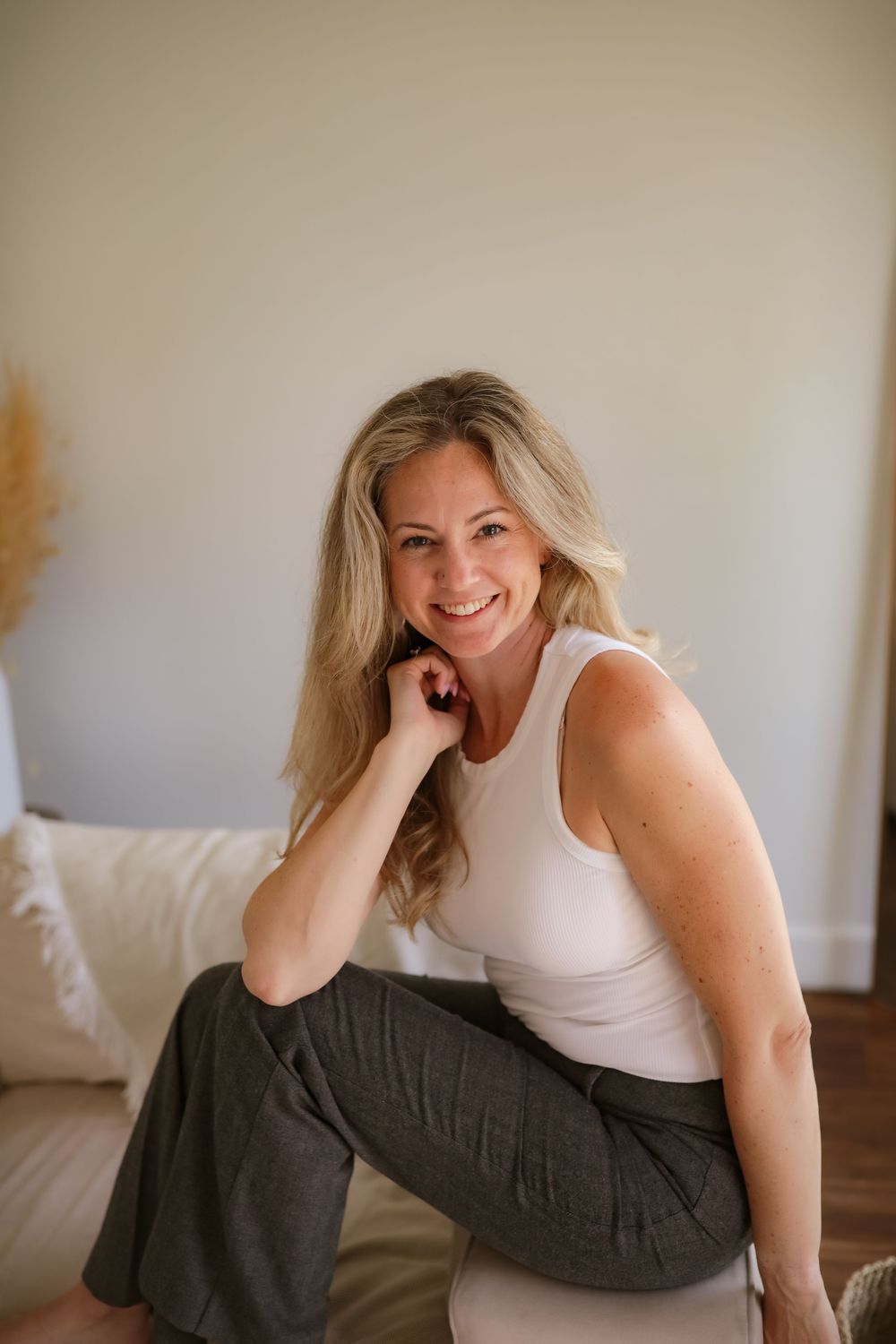 Smiling blonde woman in white tank top and olive pants sitting relaxed indoors in a bright, minimalist room.