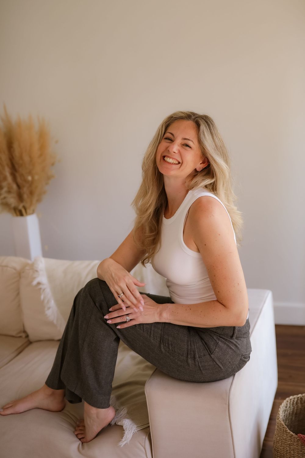 Happy blonde woman laughing while seated on a white couch wearing a white tank and gray trousers.