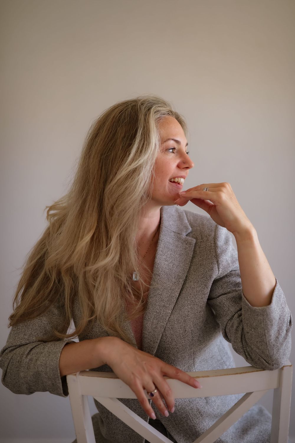 Blonde woman in gray blazer sitting on chair, hand near chin, looking sideways in bright room.