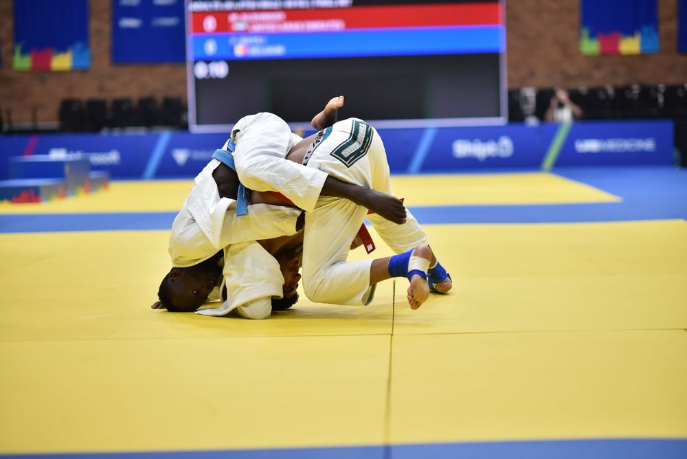 Two judo athletes compete on a yellow mat during an official judo tournament match in a sports arena.