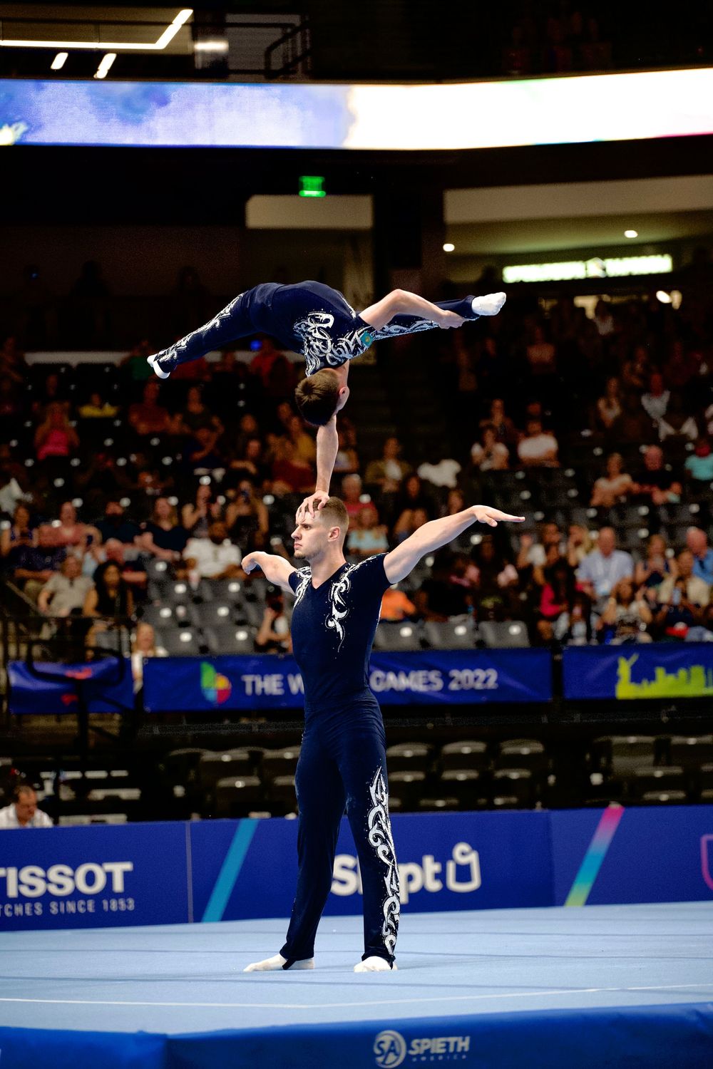 Two acrobatic gymnasts perform a balance routine at The World Games 2022, one athlete lifted overhead in a graceful pose.