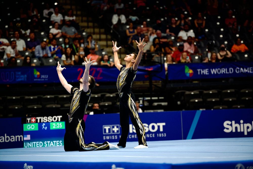 Two US acrobatic gymnasts perform a synchronized routine at The World Games in Alabama, wearing black and gold uniforms.