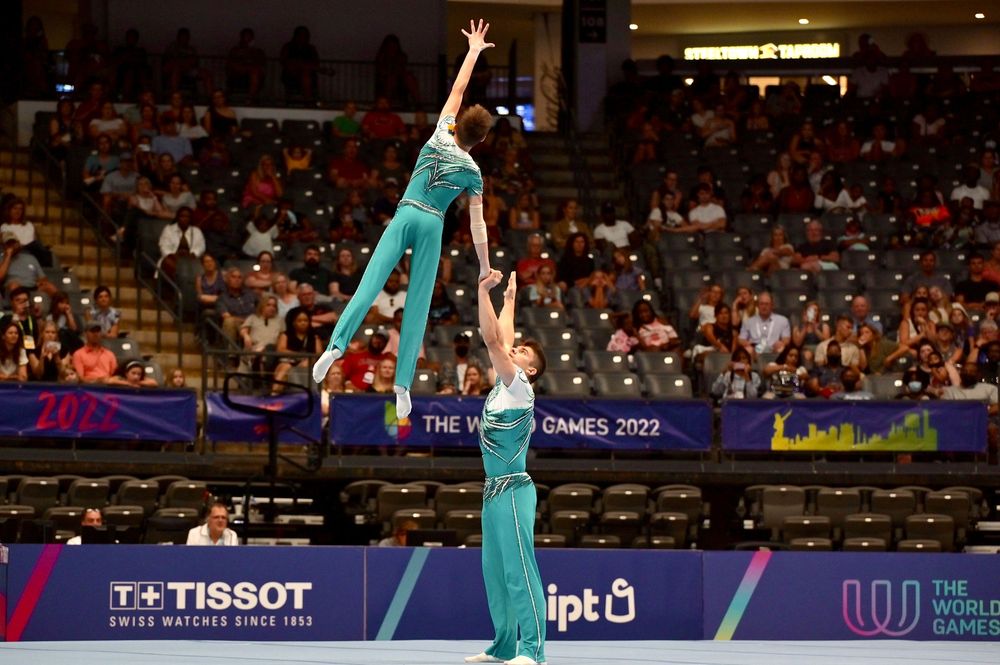 Acrobatic gymnasts in teal perform a lift at The World Games 2022 with Tissot sponsorship visible.
