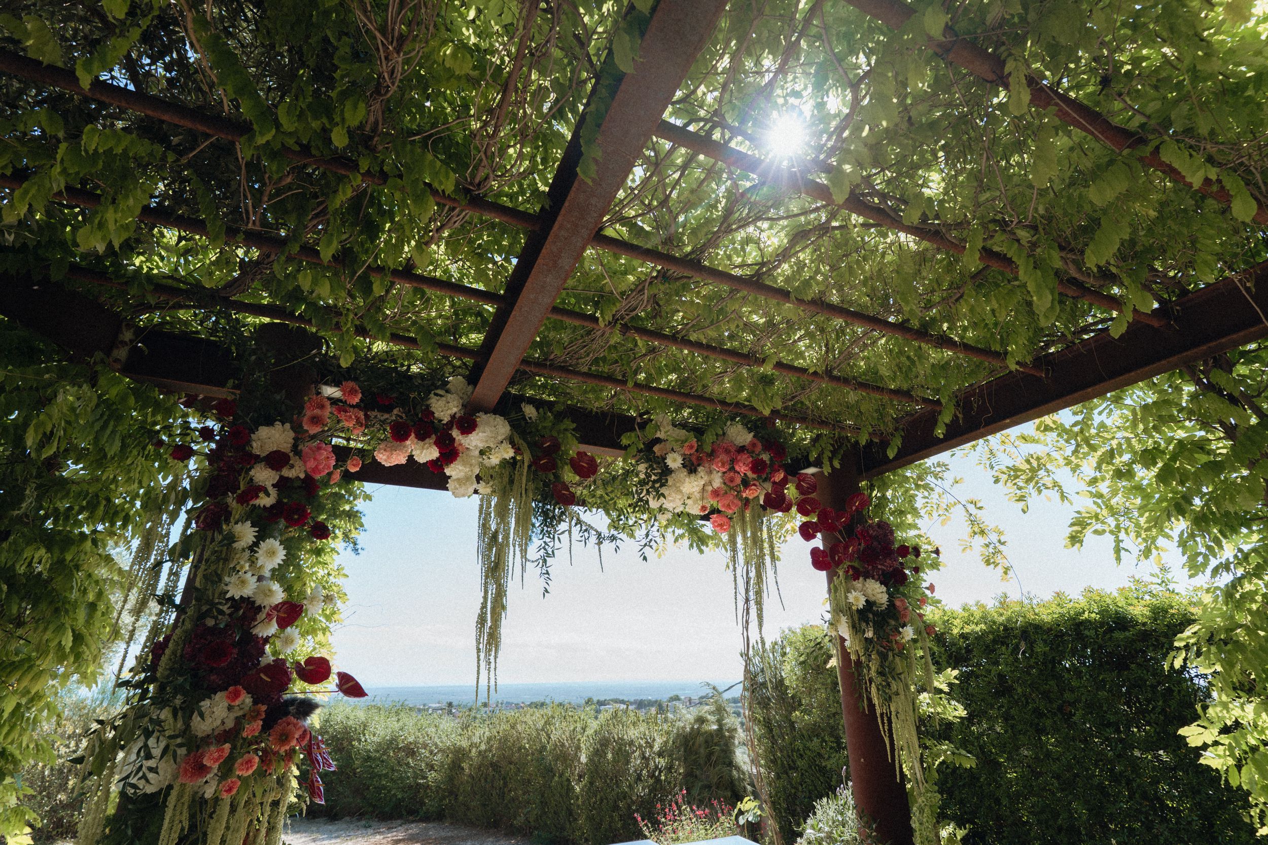 Floral wedding arch with pink and white blooms under a lush green pergola with sunlight filtering through.