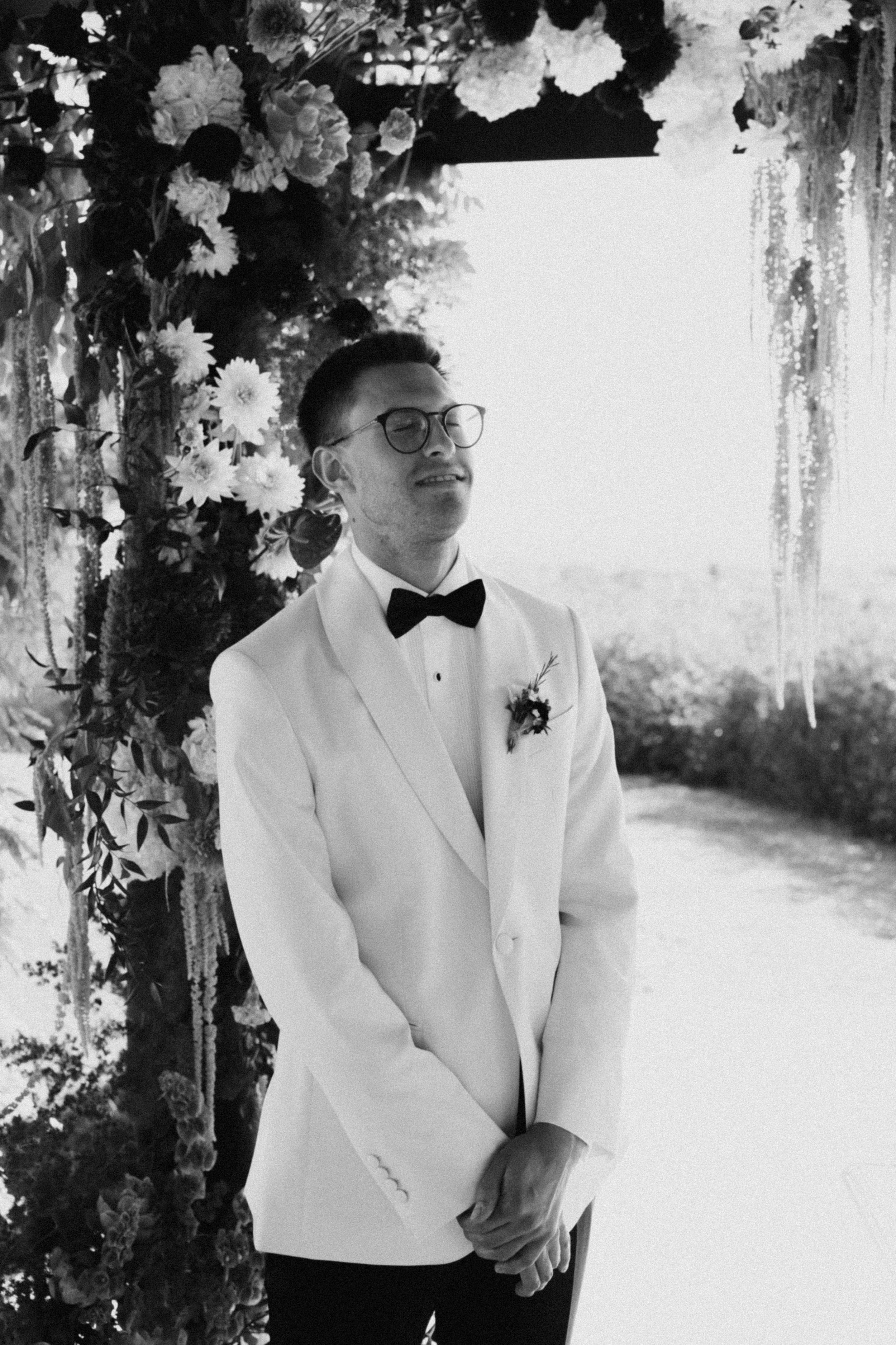 Groom in white tuxedo jacket and bow tie stands under floral arch in black and white wedding photo.