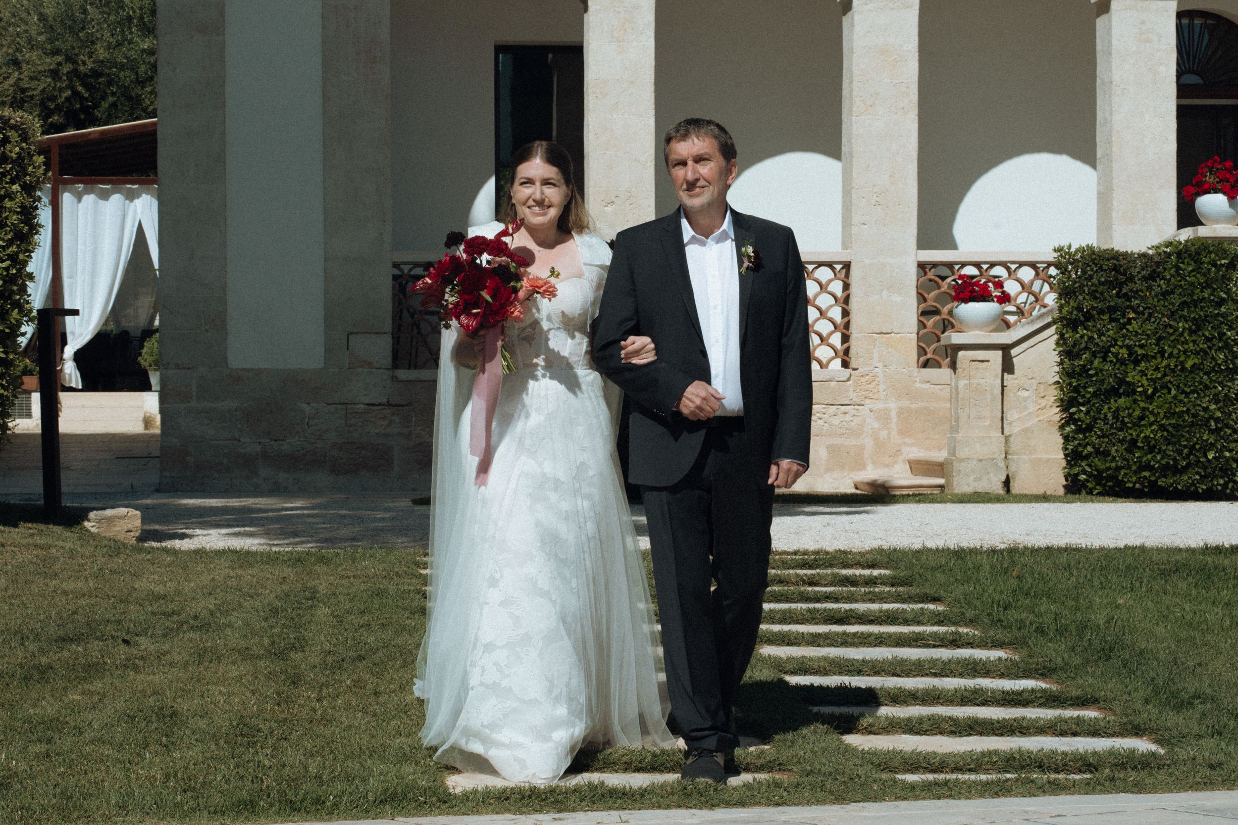 Bride in white gown holding red bouquet walks with suited man on green lawn outside elegant stone building.
