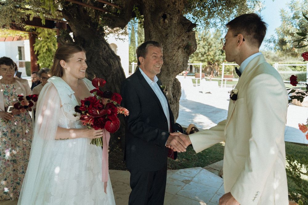 Bride in white gown holds red bouquet as groom shakes hands with officiant under a large tree at outdoor wedding.
