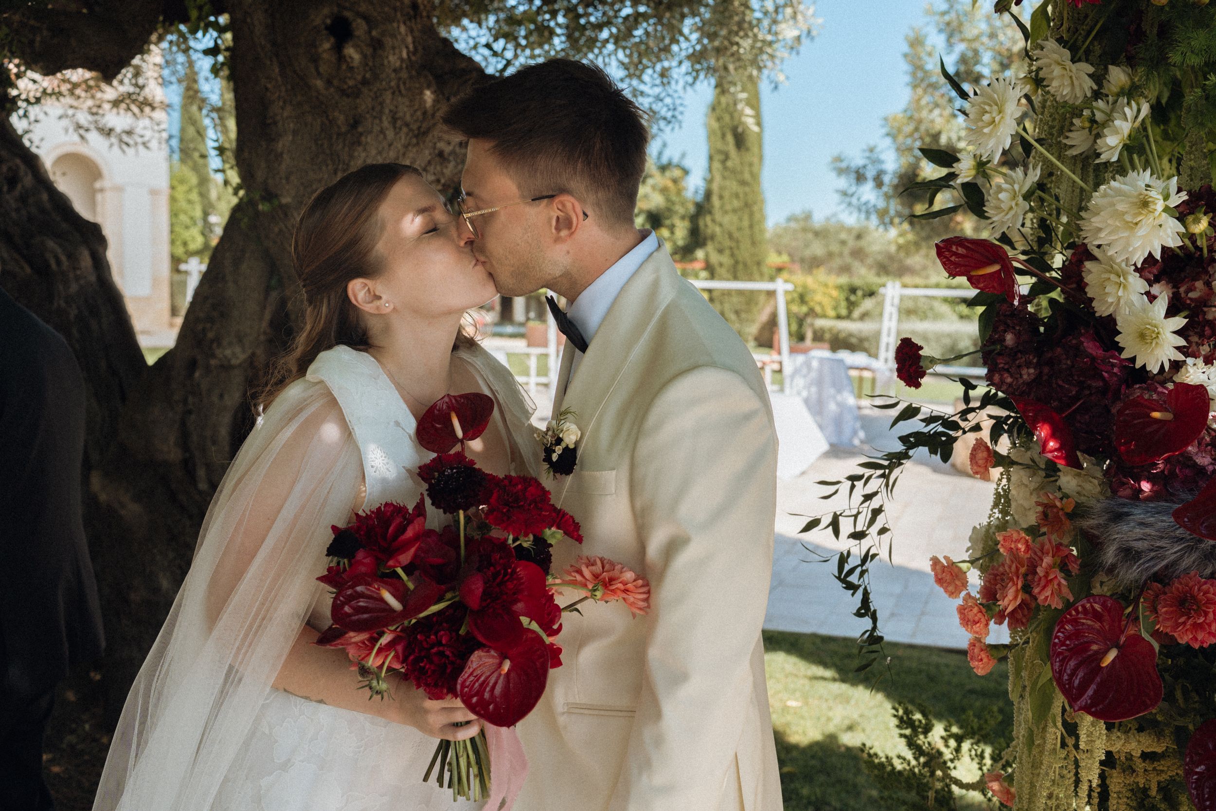 Bride and groom share a romantic kiss outdoors at a wedding ceremony with red floral bouquet and white blooms.