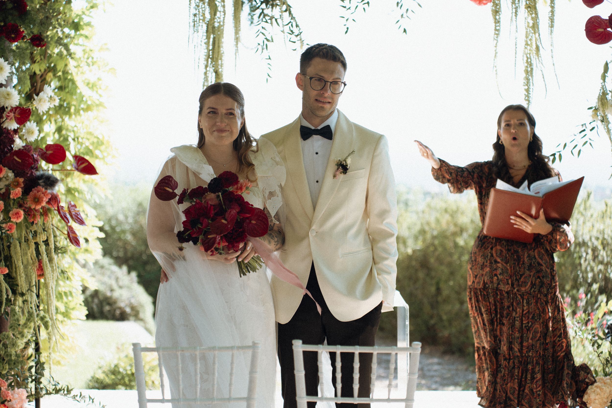 Bride and groom stand at outdoor wedding altar with red floral arrangements as officiant reads ceremony.