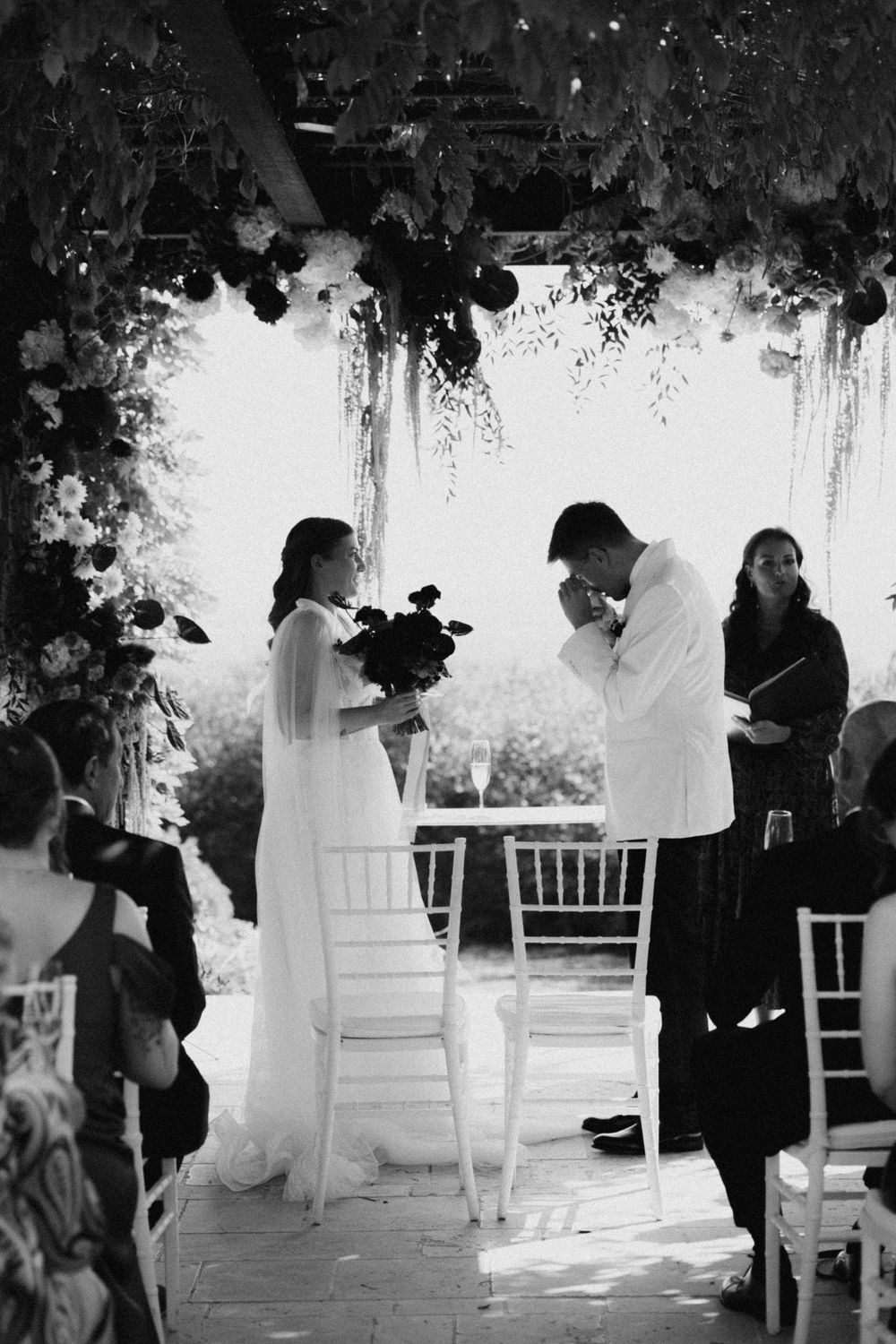 Black and white wedding ceremony photo of bride and groom exchanging vows under floral arch with guests seated nearby.