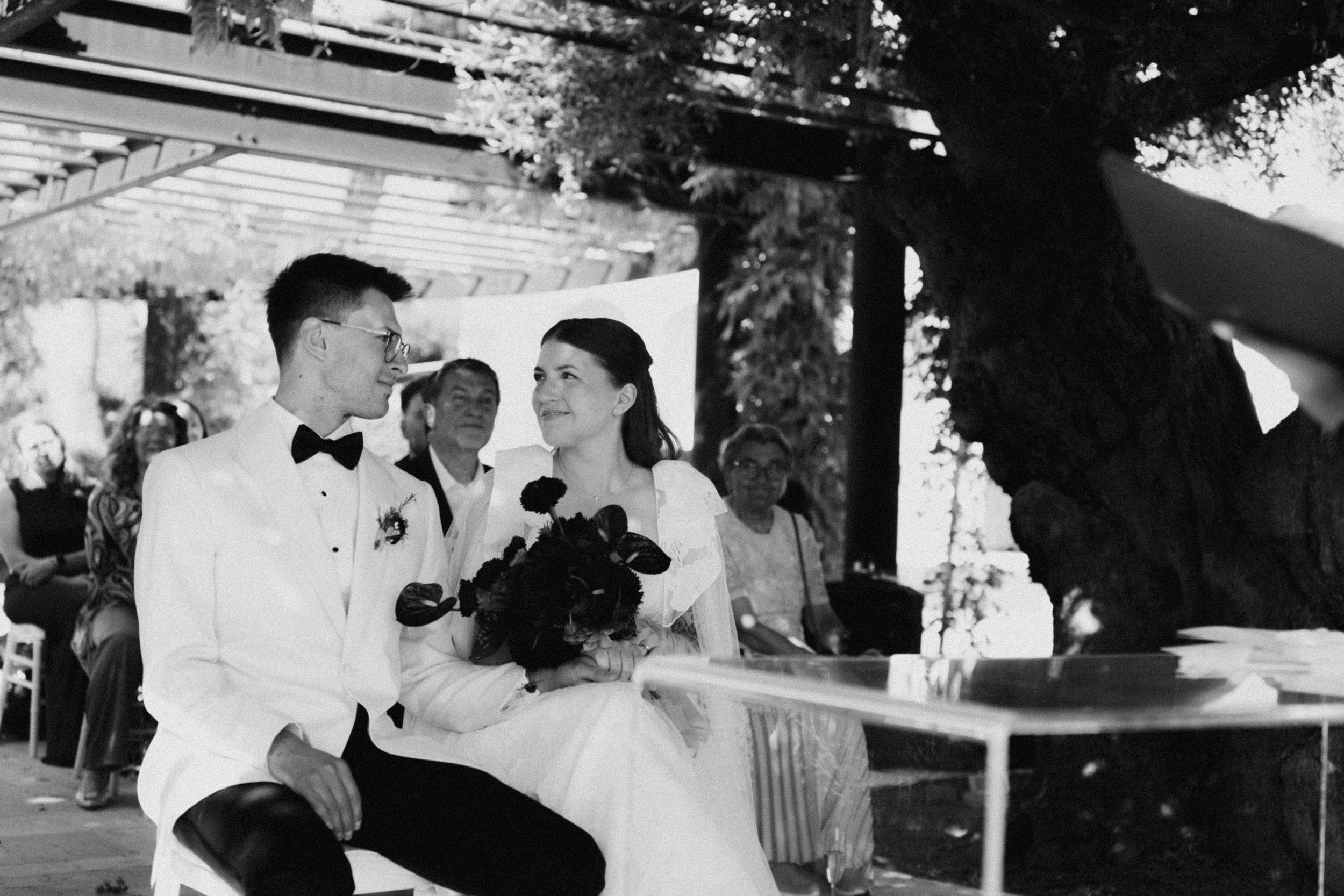 Black and white photo of a smiling bride and groom sitting together during their outdoor wedding ceremony.