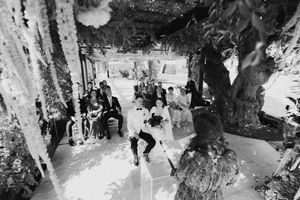 Black and white wedding ceremony photo with guests seated outdoors under floral decorations and trees.