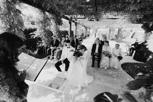 Black and white outdoor wedding ceremony with bride, groom, and guests gathered under a tree-covered pergola.
