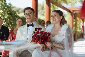 Smiling bride holding red roses bouquet and groom in white tuxedo laughing during outdoor wedding ceremony.