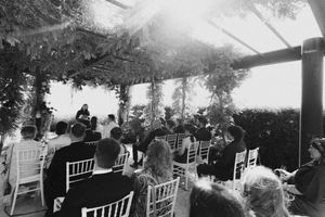 Black and white outdoor wedding ceremony with guests seated on white chairs under a vine-covered pergola.