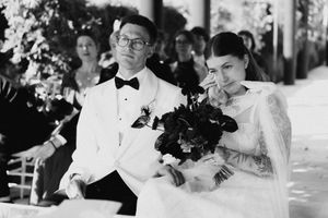 Black and white wedding photo of bride and groom seated during ceremony, bride holding bouquet and wiping tears.