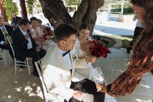 Bride and groom seated at outdoor wedding ceremony, sharing a joyful moment with a guest holding champagne glasses.