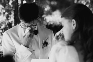 Emotional groom in white tuxedo wiping tears during wedding vows ceremony, black and white photo.
