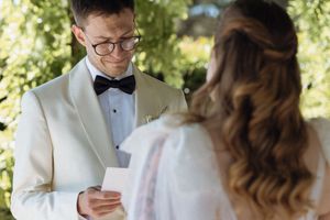 Groom in white tuxedo jacket and bow tie reads wedding vows from a card to his bride outdoors.