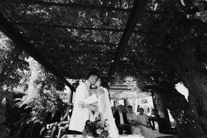 Black and white photo of a couple sharing a kiss at their outdoor wedding ceremony under a lush vine-covered pergola.