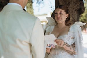 Emotional bride reading wedding vows while holding a card outdoors, wearing a white dress with bow details.