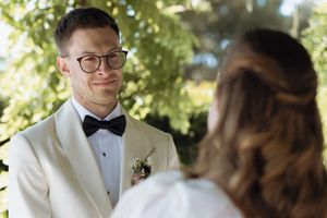 Groom in white tuxedo and bow tie smiling at bride during outdoor wedding ceremony.