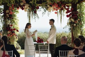 Couple exchanging vows under floral arch with red and white flowers at outdoor wedding ceremony.