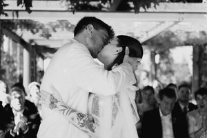 Black and white photo of groom kissing bride on forehead during outdoor wedding ceremony with guests watching.
