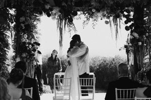 Black and white photo of newlyweds embracing under floral arch at outdoor wedding ceremony with seated guests.