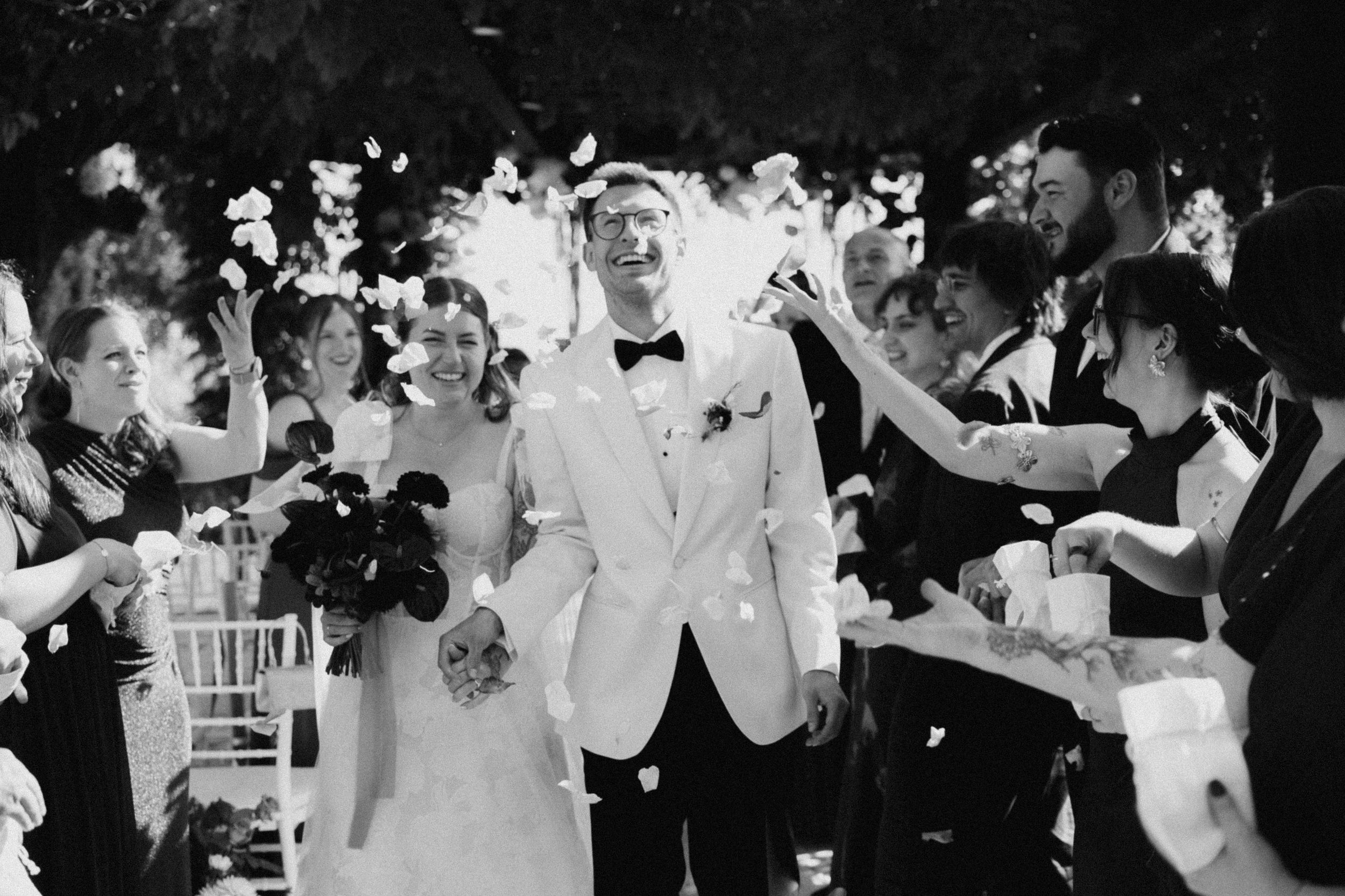 Black and white photo of a bride and groom walking down the aisle as guests toss confetti at their outdoor wedding.