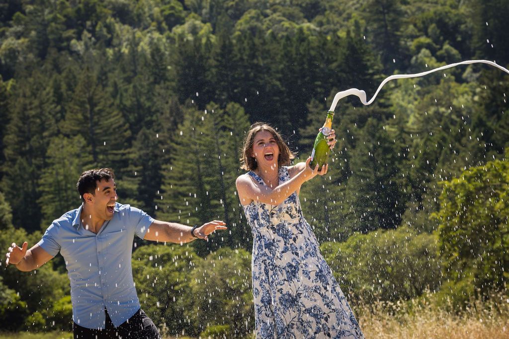 Joyful couple celebrating outdoors, woman in floral dress spraying champagne bottle with green forest background.