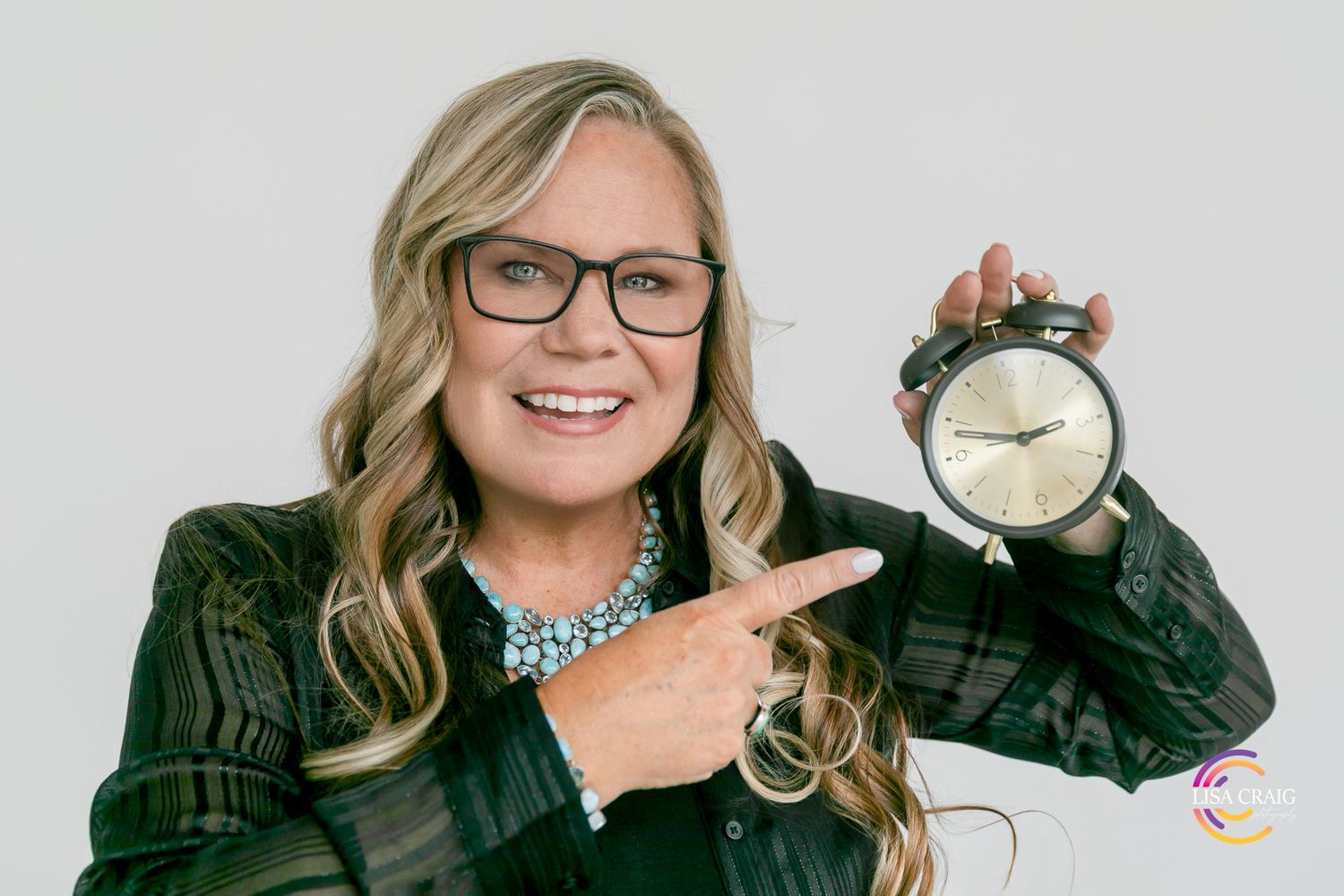 Smiling woman with glasses and blonde hair pointing at a vintage alarm clock against a white background.