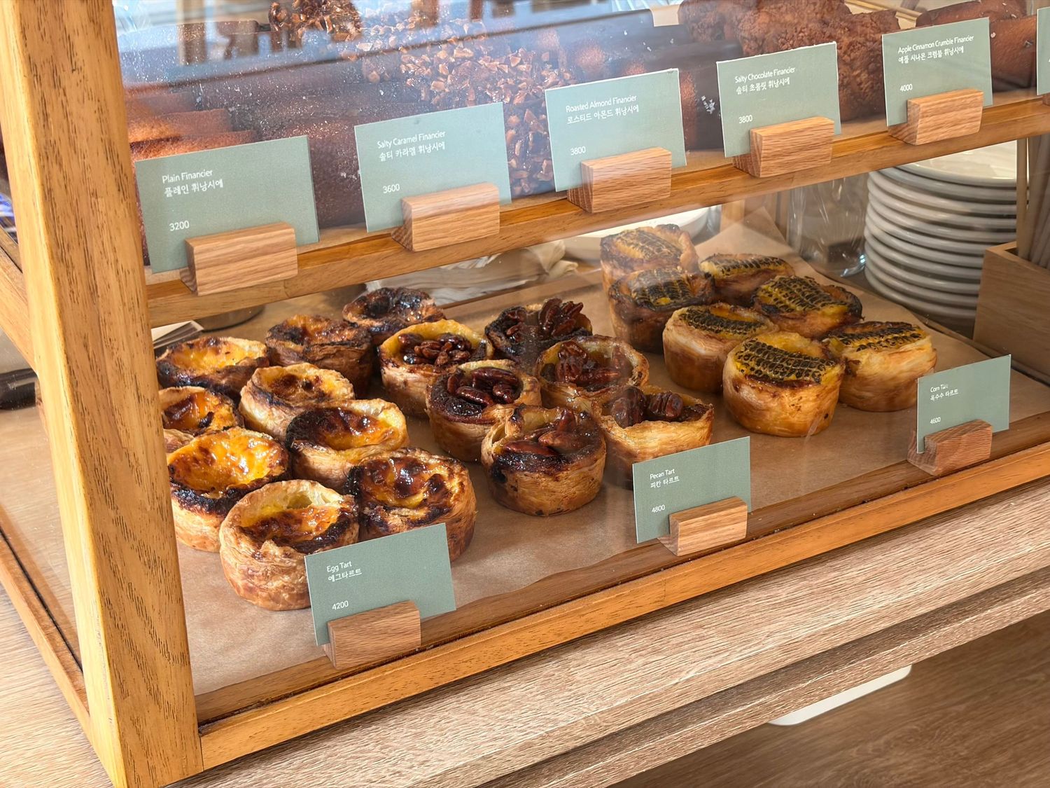 Wooden bakery display case filled with assorted Portuguese custard tarts and fruit pastries with price tags.