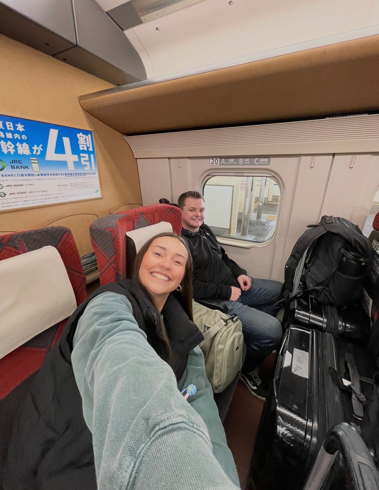 Two travelers smile for a selfie on a Japanese Shinkansen bullet train, surrounded by luggage and red seats.
